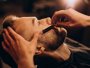 Há na imagem um homem sentado em uma cadeira. Uma 
                mão segurando uma navalha se aproxima da barba do homem.